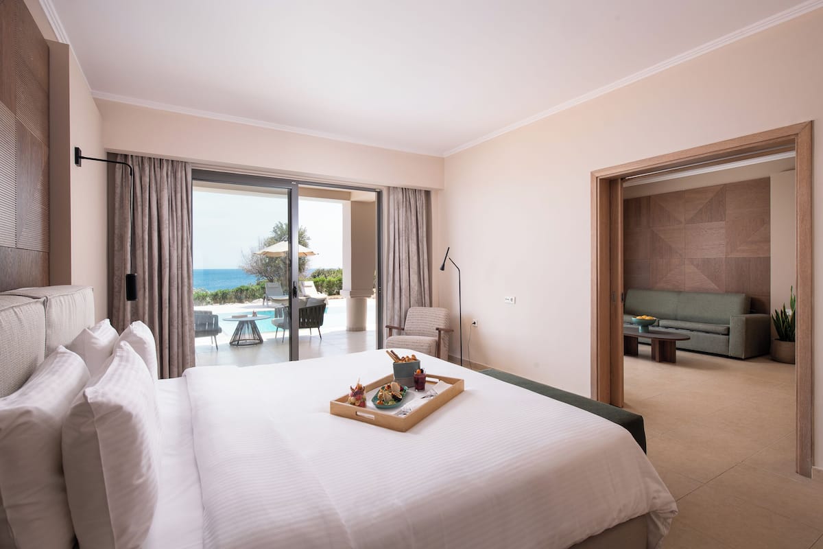 Quarto de hotel com uma cama grande, bandeja de itens de café da manhã, varanda com vista para o mar e área de estar com sofá e mesa.