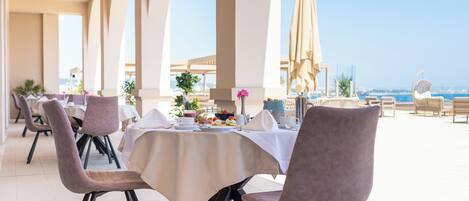Un espace repas avec des tables dressées pour le repas, des chaises et une vue sur la mer.