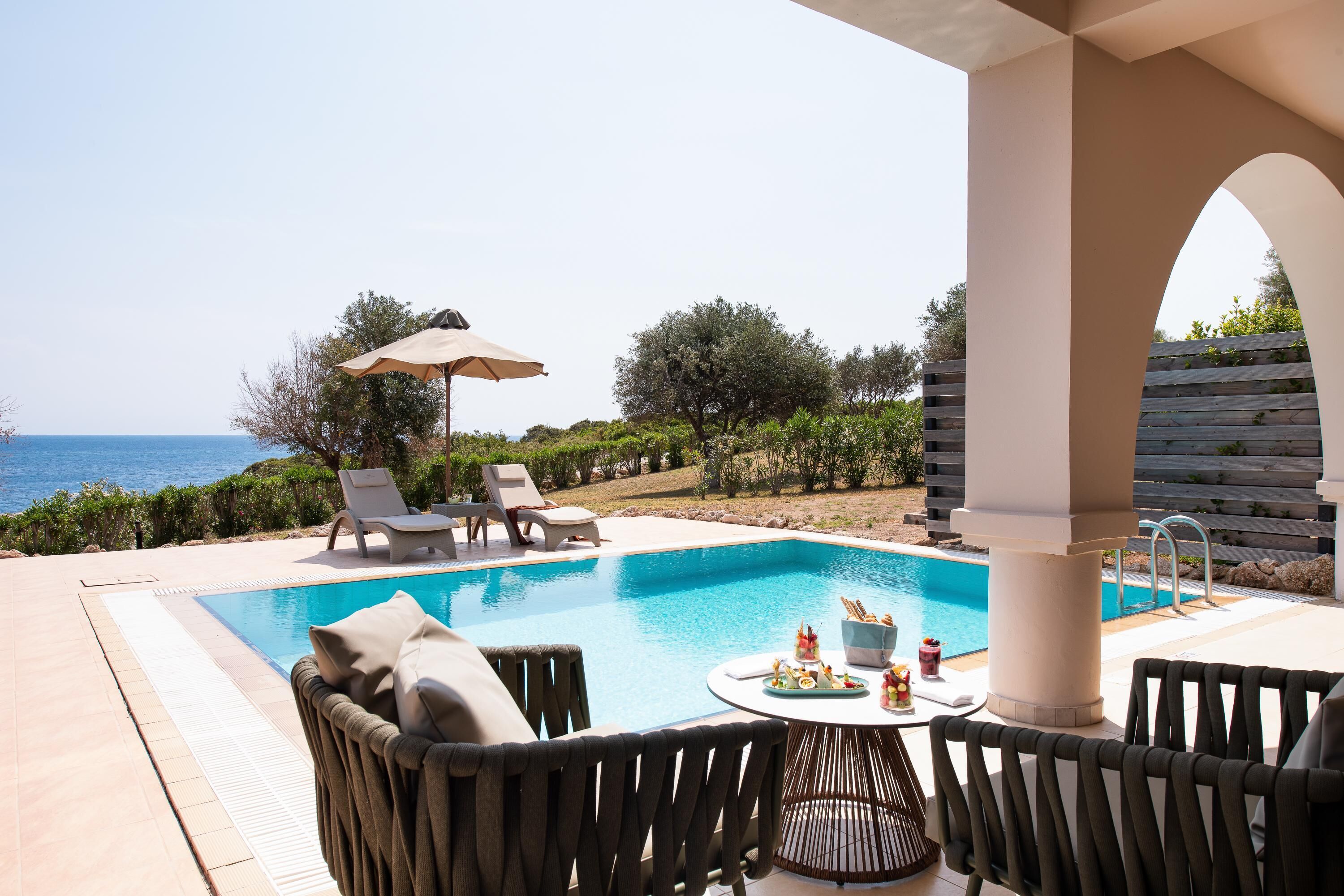 Un área junto a la piscina con sillones de descanso, una mesa con aperitivos y vistas al mar.
