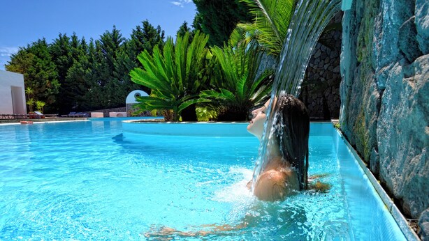 A swimming pool with clear blue water, surrounded by greenery and a stone wall. - Gattopardo Park Hotel (Lipari)