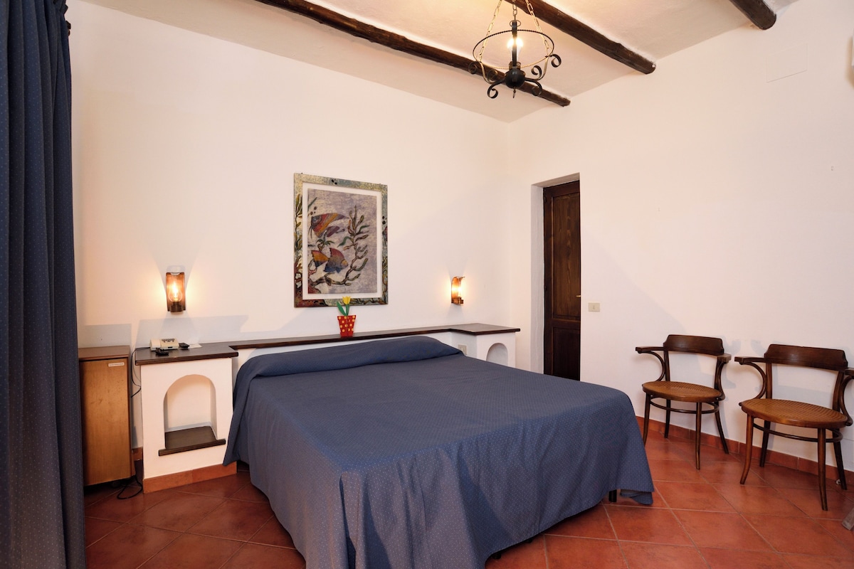 A bedroom with a bed, a desk, two chairs, a small cabinet, and a painting on the wall.