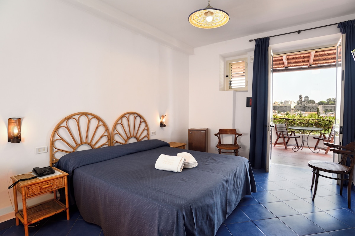 A hotel room with a bed, bedside table, lamp, and a view of the outside balcony.
