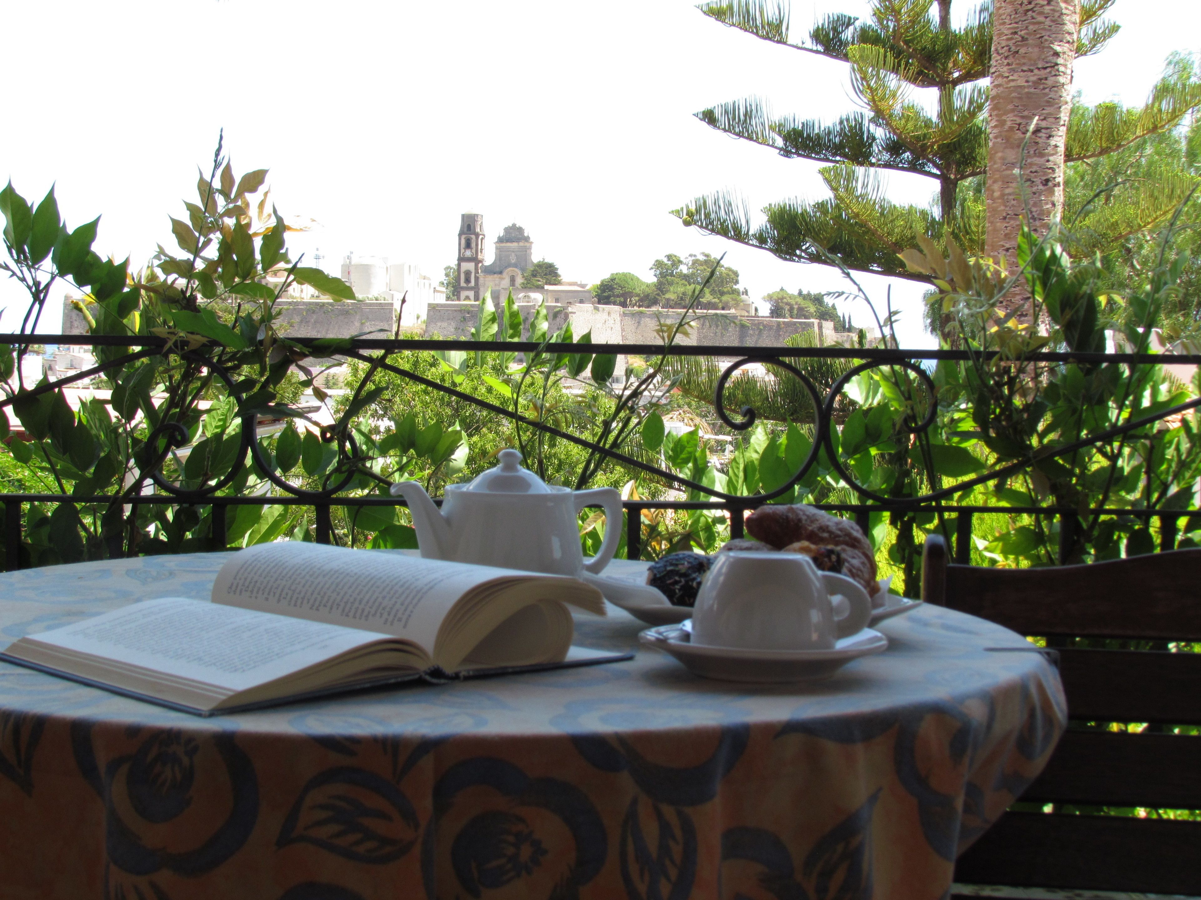 Una mesa con un libro, tetera, tazas y pasteles en un balcón con vistas a un paisaje urbano.