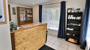 A reception area with a wooden desk, a glass display cabinet, a window with curtains, and a shelf with various items. - Motel Des Deux Lacs Inc (Shefford)