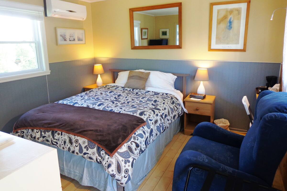 Ein Hotelzimmer mit einem Bett, zwei Nachttischen mit Lampen, einem Sessel, einem Fenster und Wandbildern.