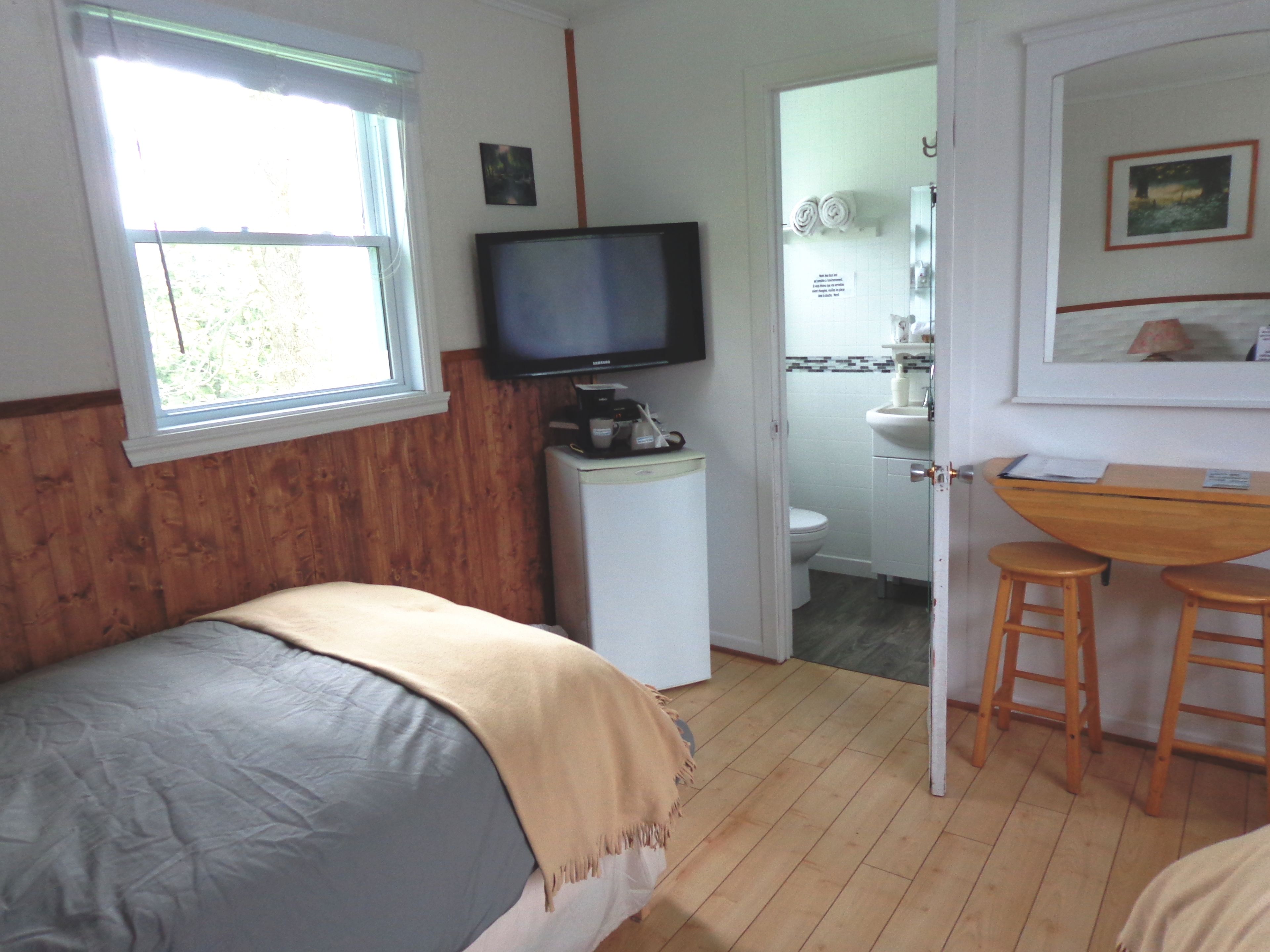 A room with a bed, a television on a small table, a wooden desk with two stools, and a bathroom visible through an open door.
