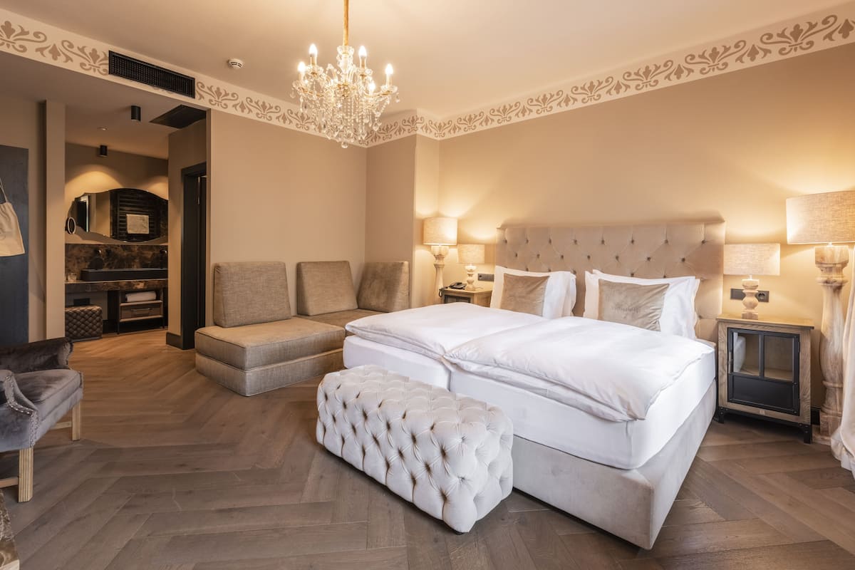 Um quarto espaçoso com uma cama grande, um espaço de estar com sofá e banquinho, um lustre e uma mesa de cabeceira com luminárias.