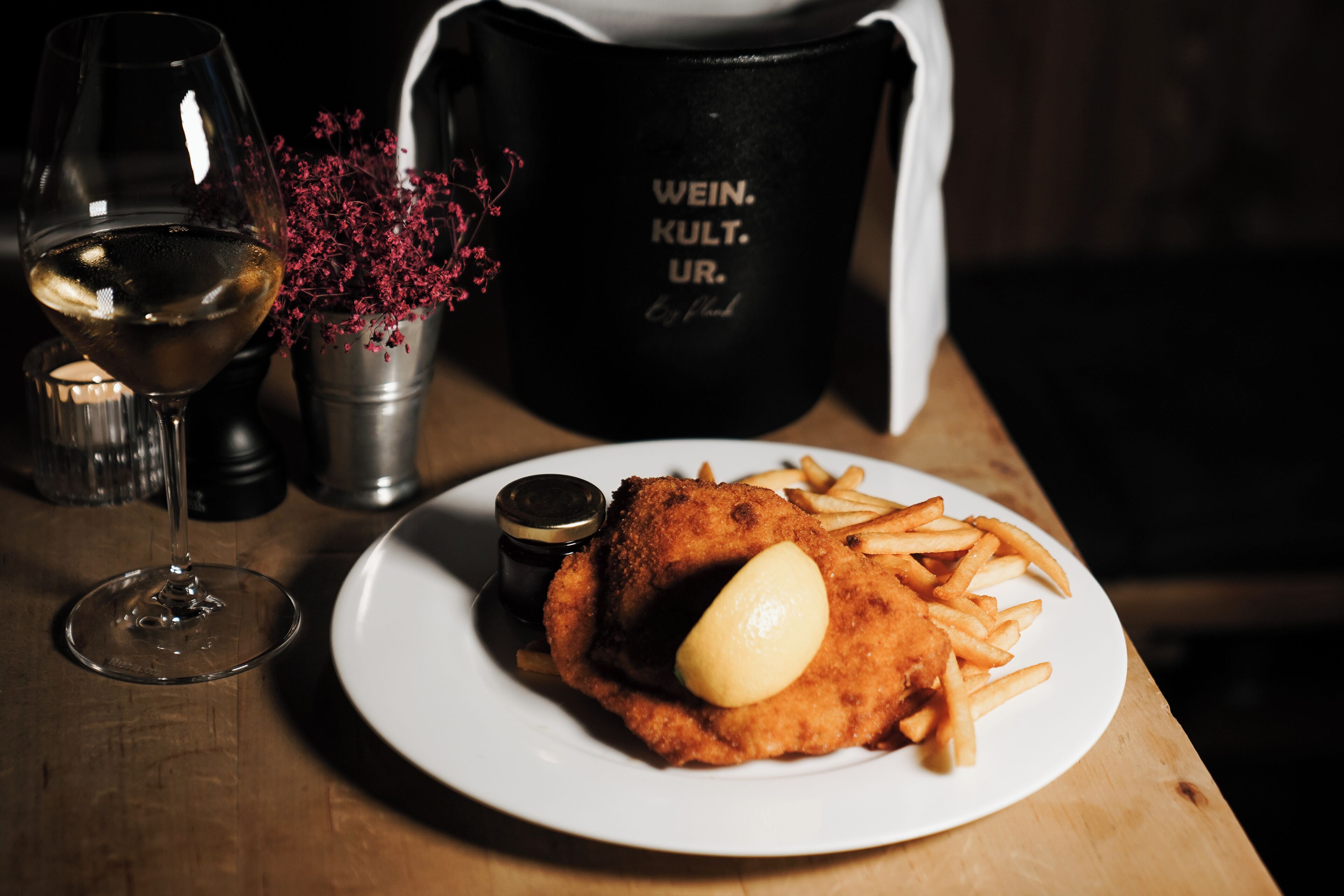 A plate of fried fish and fries with a lemon wedge, a glass of white wine, and a black mug with text on a wooden table.