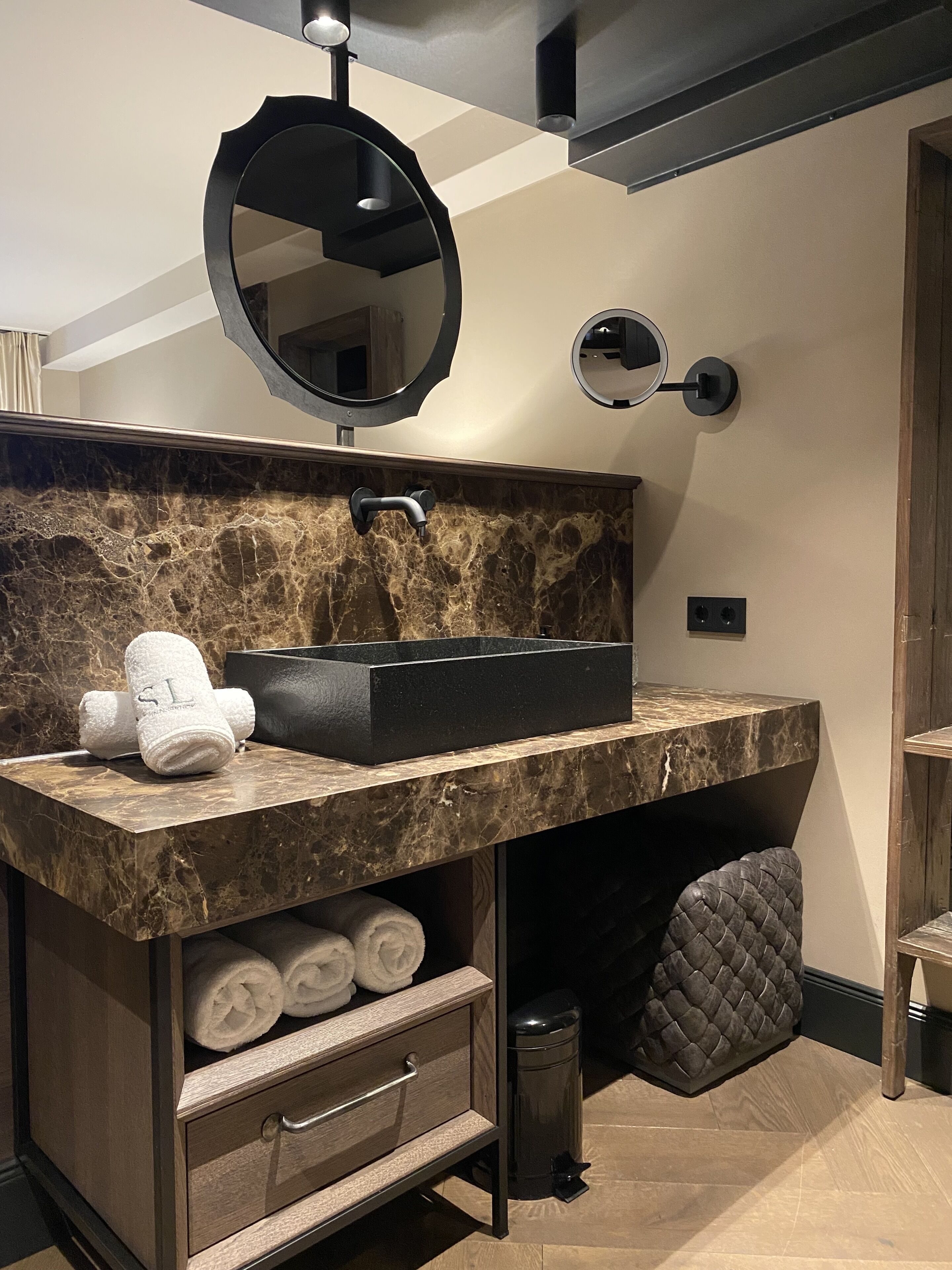 Un baño moderno con encimera de mármol, un lavabo negro, un espejo redondo y un mueble para toallas.