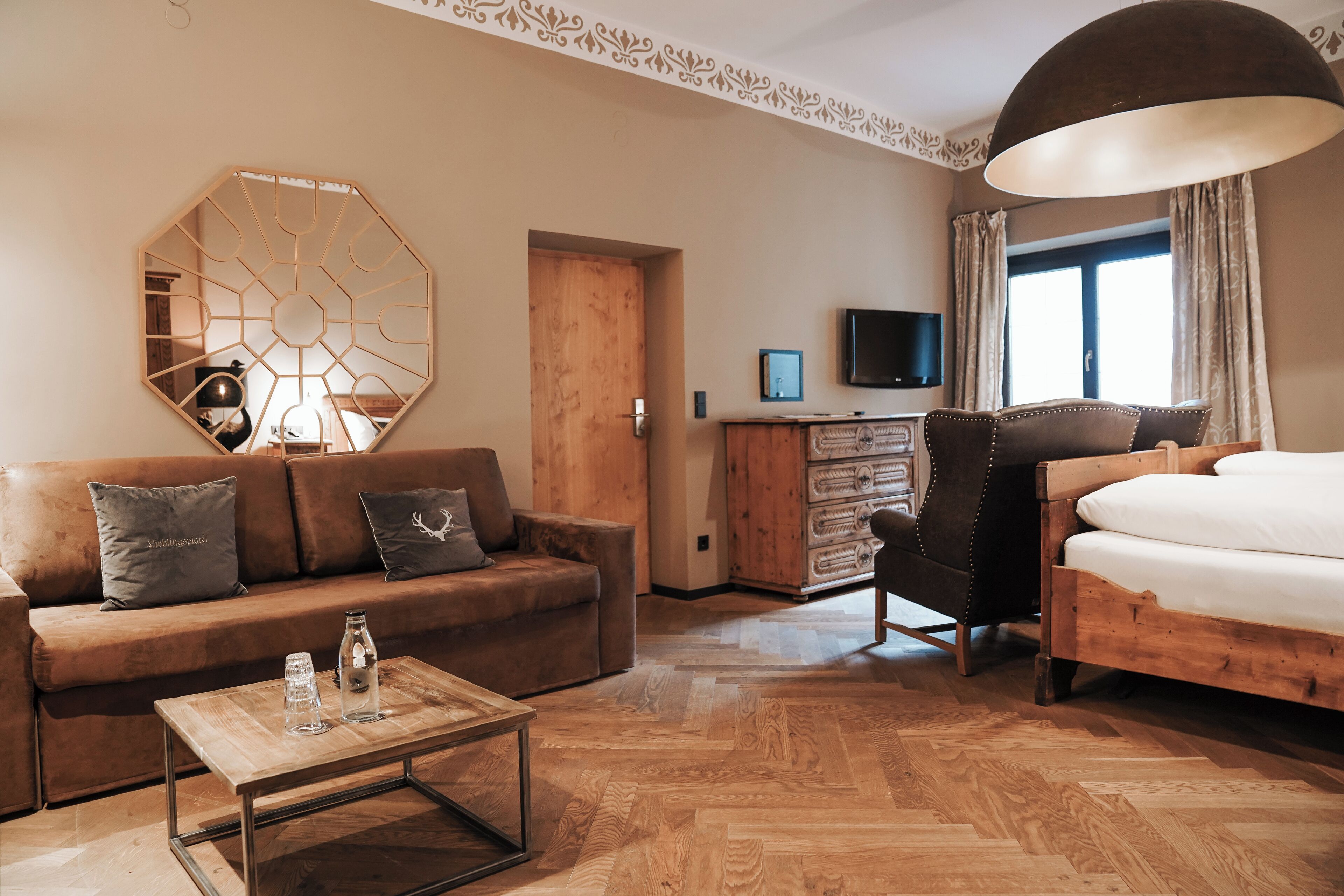 A hotel room with a wooden herringbone floor, a sofa, a bed, a desk with a TV, and a large window with curtains.