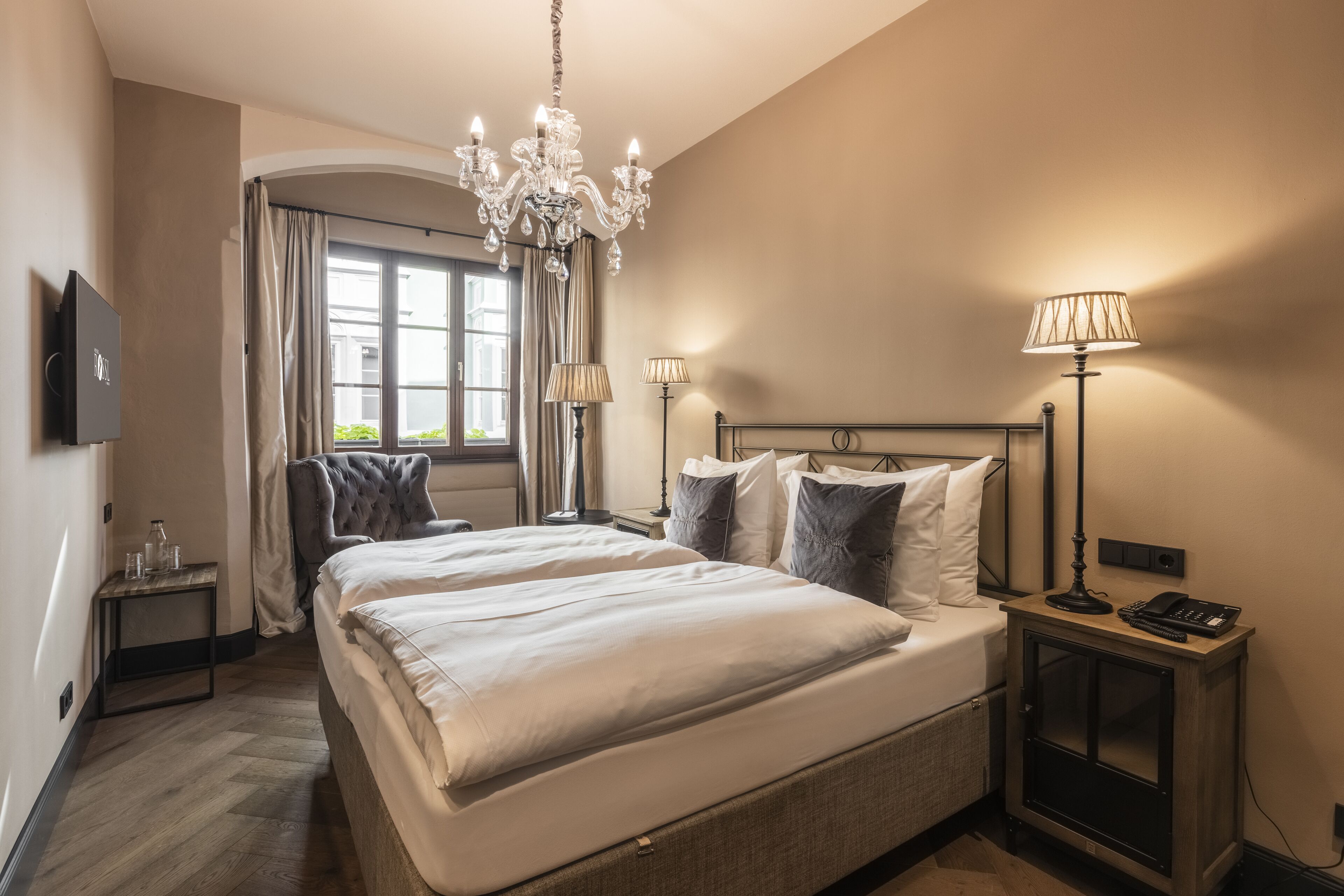 Habitación de hotel con una cama grande, dos lámparas de noche, un candelabro, un televisor y una mesita con un teléfono y una lámpara.