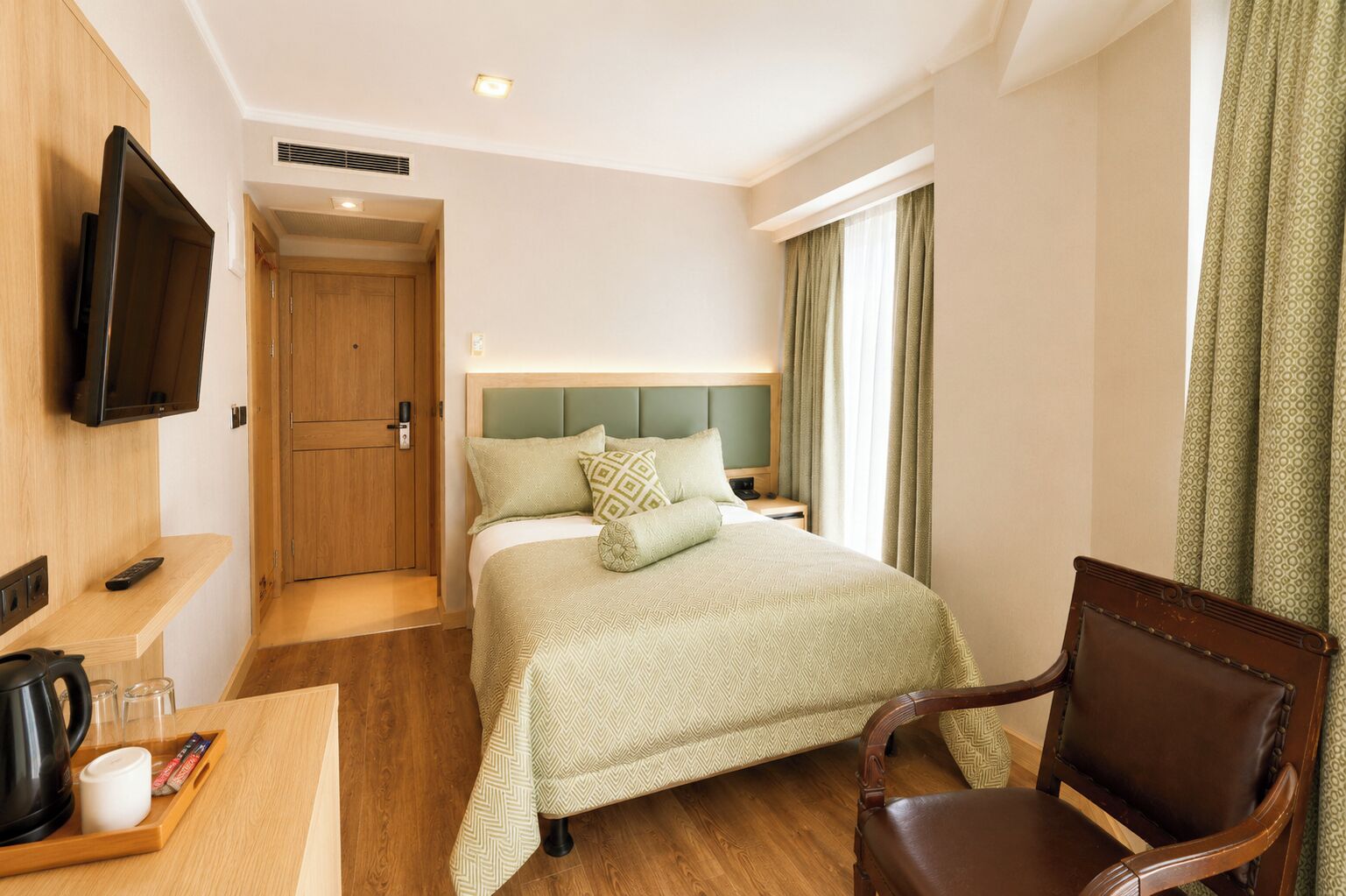 Habitación de hotel con cama, televisor montado en la pared, escritorio de madera con tetera y vasos, y una silla.