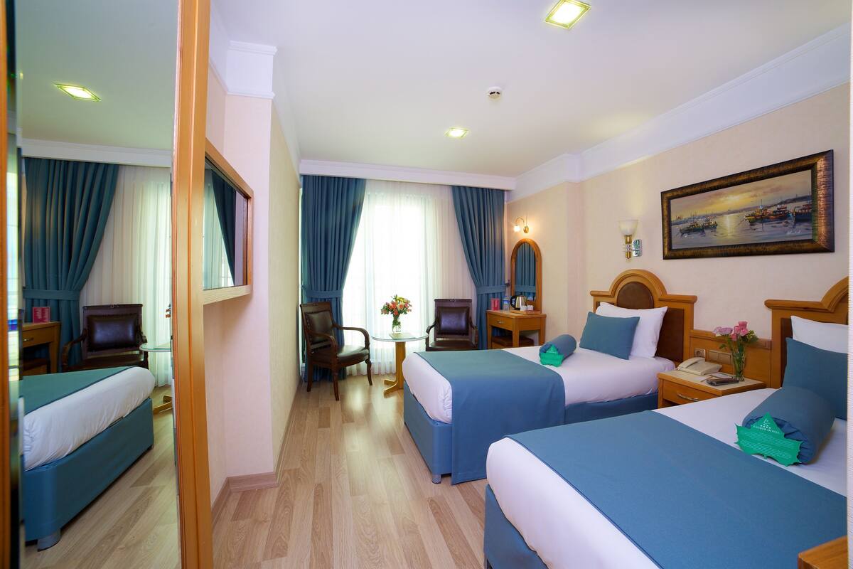 A hotel room with two beds, a desk, a chair, a small table with a vase, and a painting on the wall.