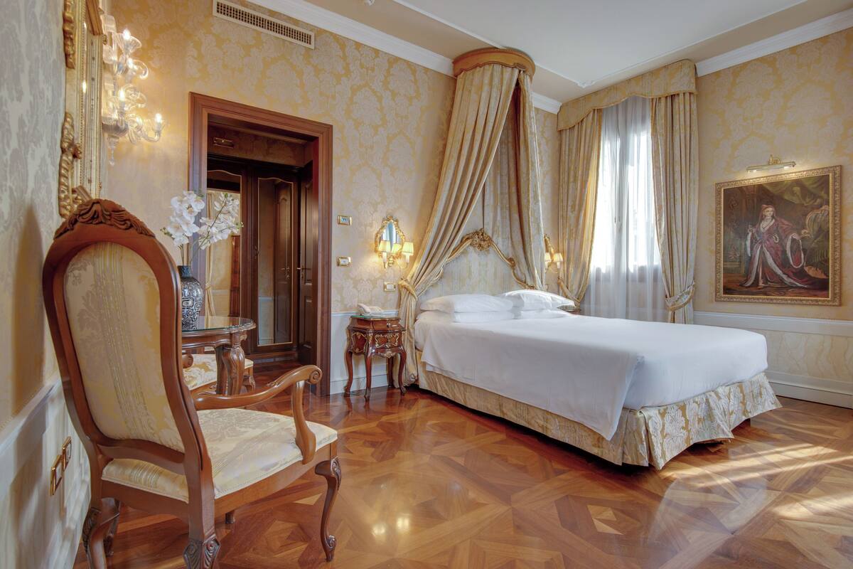 A bedroom with a large bed, a sitting area with a chair, a small table, and a mirror.
