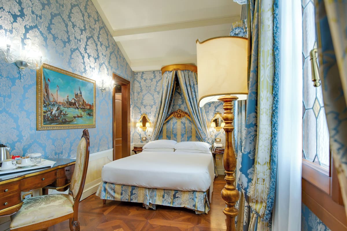 A bedroom with a bed, a desk, a chair, a nightstand, a lamp, and a painting on the wall.