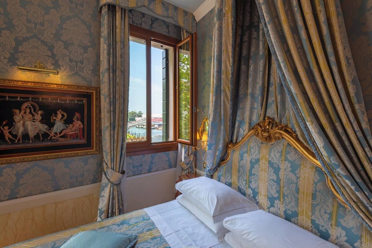 A bedroom with a bed, a window with curtains, a painting on the wall, and a view of a bridge and water outside.