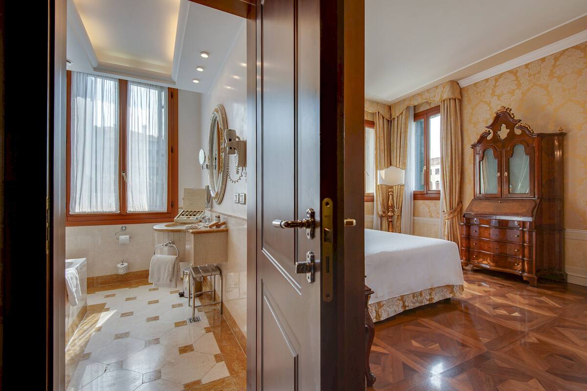 A hotel room with a bed, a vanity with a mirror, a wooden dresser, and a bathroom with a toilet and sink.