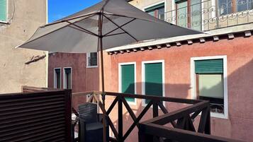 Un balcón con una sombrilla grande, muebles de mimbre y un edificio con contraventanas verdes.
