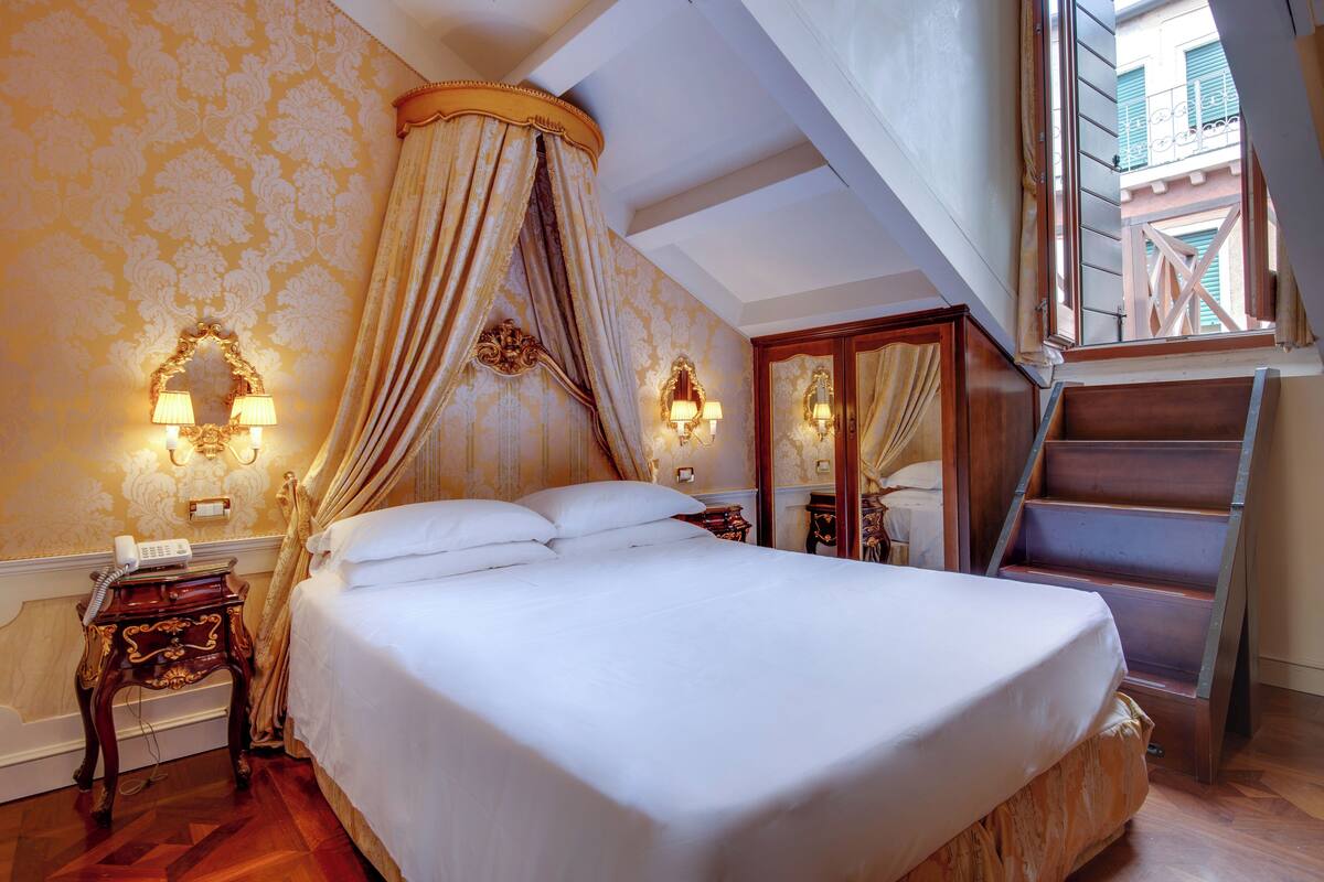 A bedroom with a large bed, a canopy, two wall sconces, a telephone, and a wooden ladder.