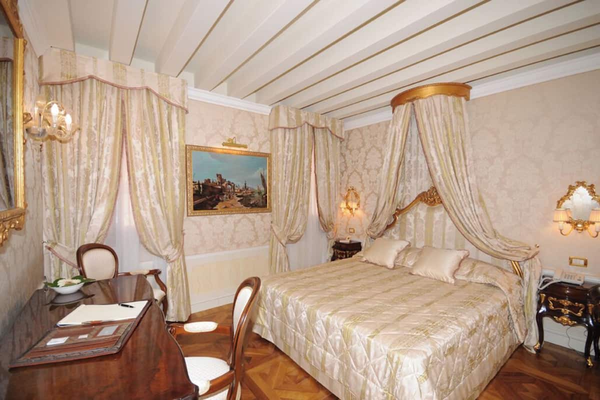 A bedroom with a four-poster bed, a desk with a chair, a nightstand, and a painting on the wall.