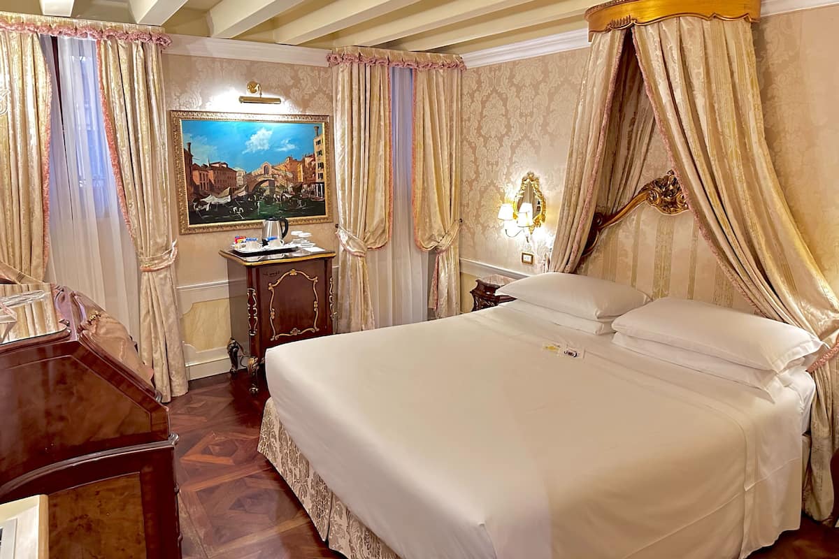 A hotel room with a large bed, a desk with a lamp, a painting on the wall, and ornate curtains.