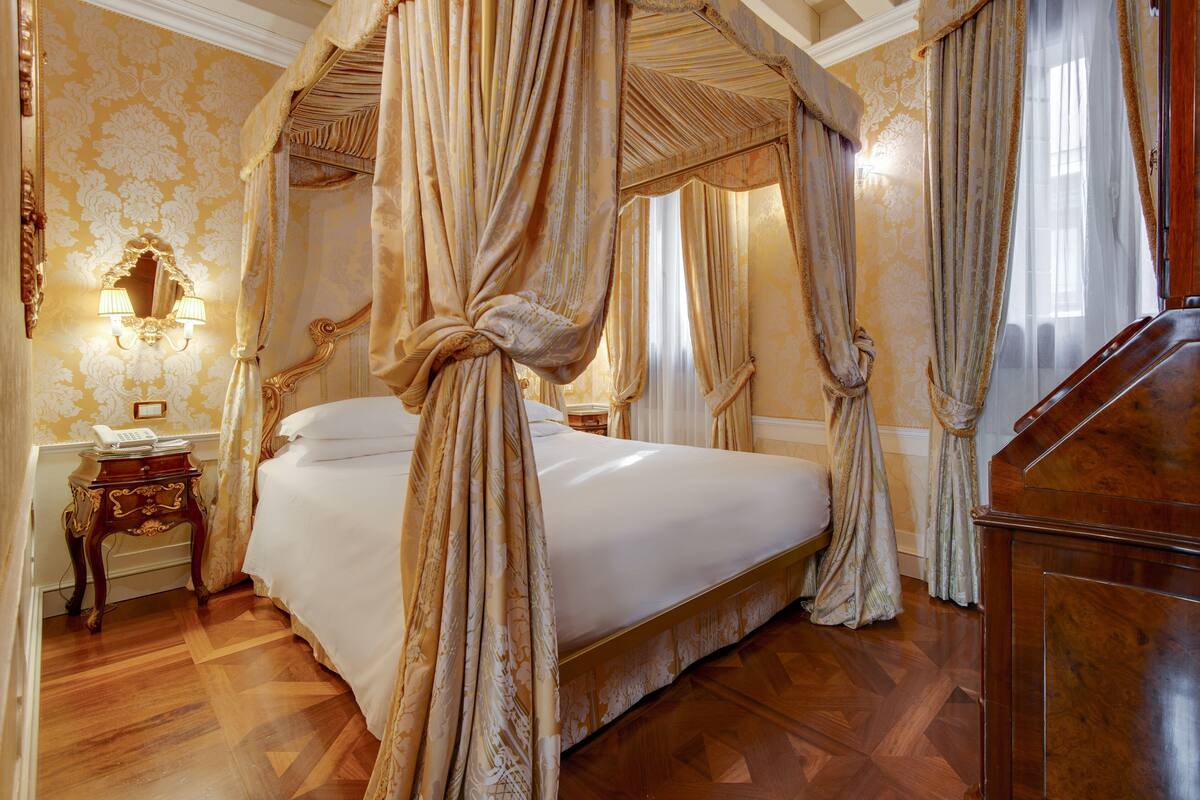 A four-poster bed with a canopy, a wooden nightstand with a telephone, and a mirror with wall sconces.