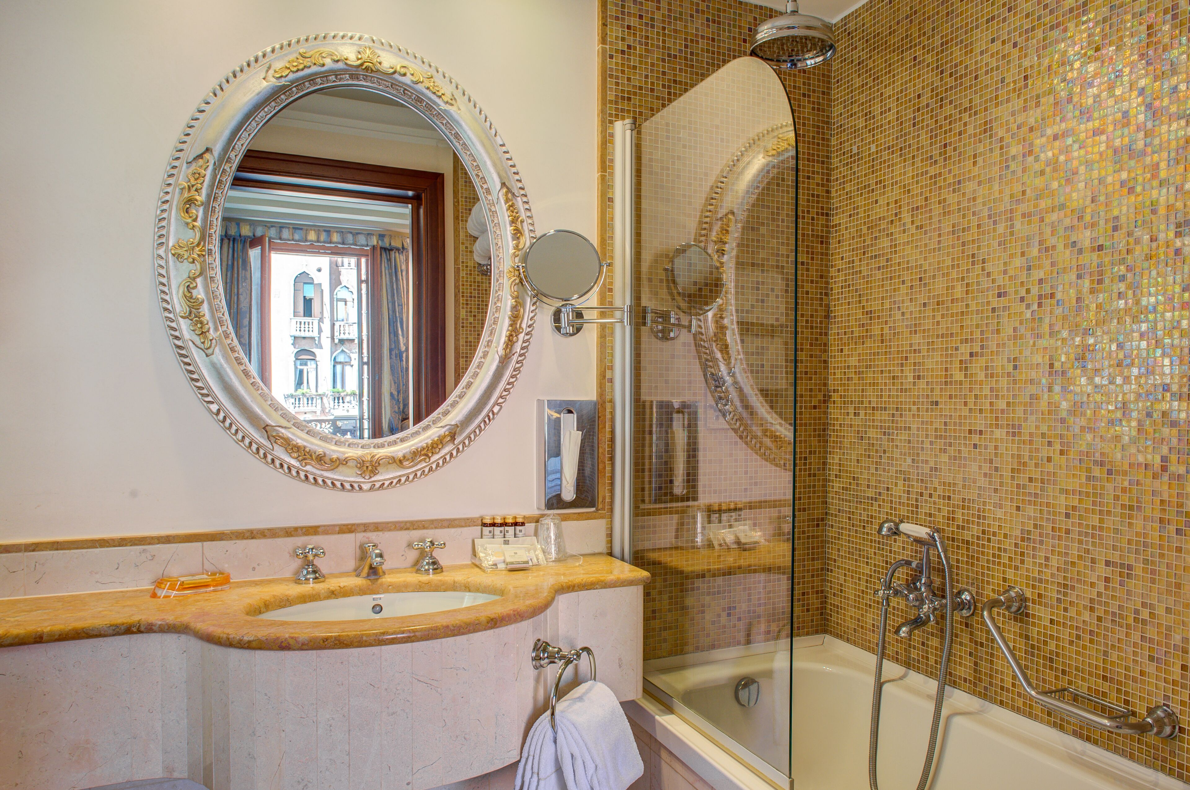 A bathroom with a large ornate mirror, a sink with two faucets, a glass-enclosed shower, and a mosaic-tiled wall.