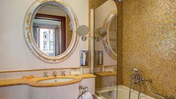 A bathroom with a large ornate mirror, a sink with two faucets, a glass-enclosed shower, and a mosaic-tiled wall.