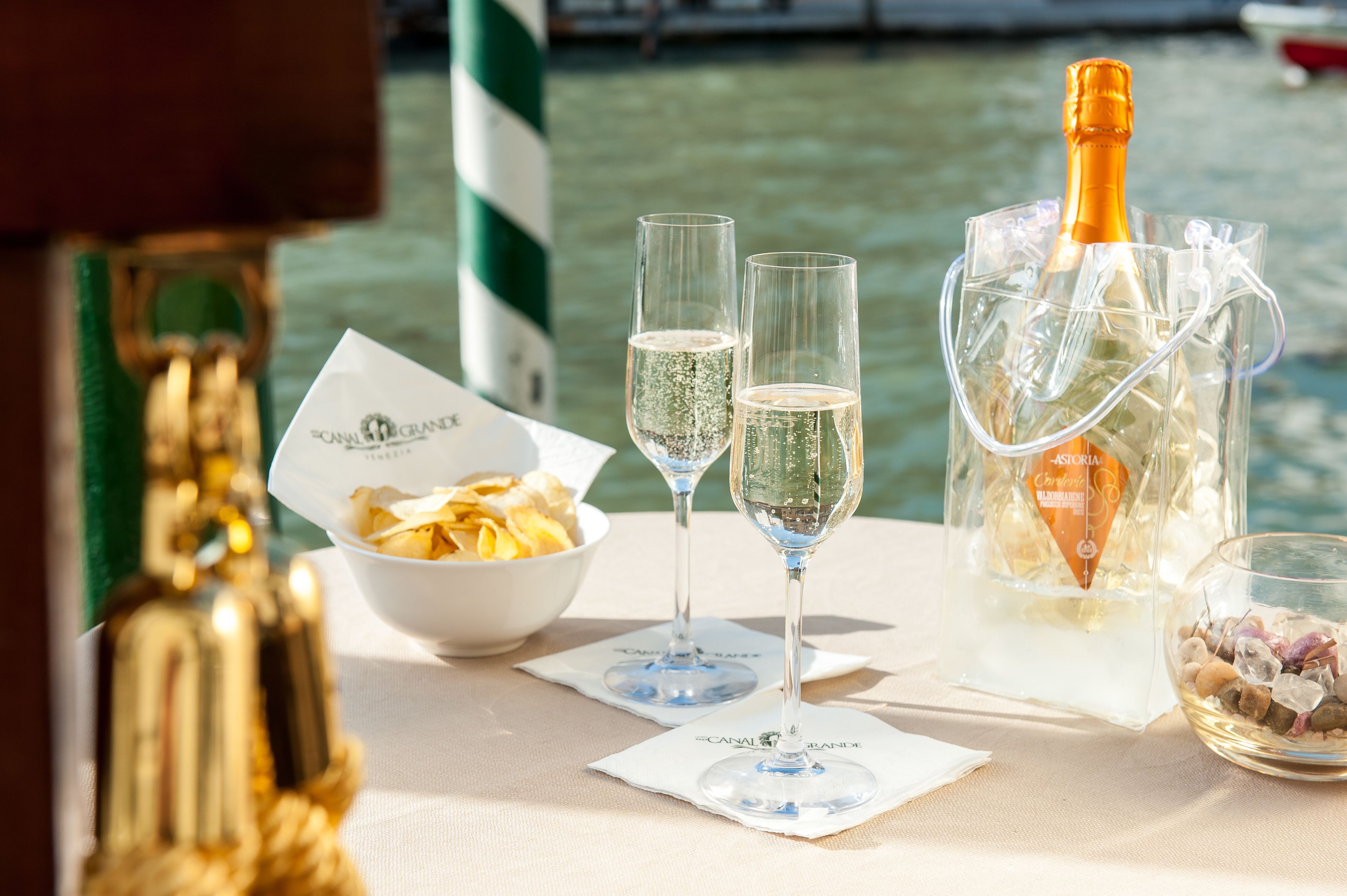 A table with two champagne glasses, a bottle of champagne in a clear ice bucket, a bowl of snacks, and a decorative bottle.