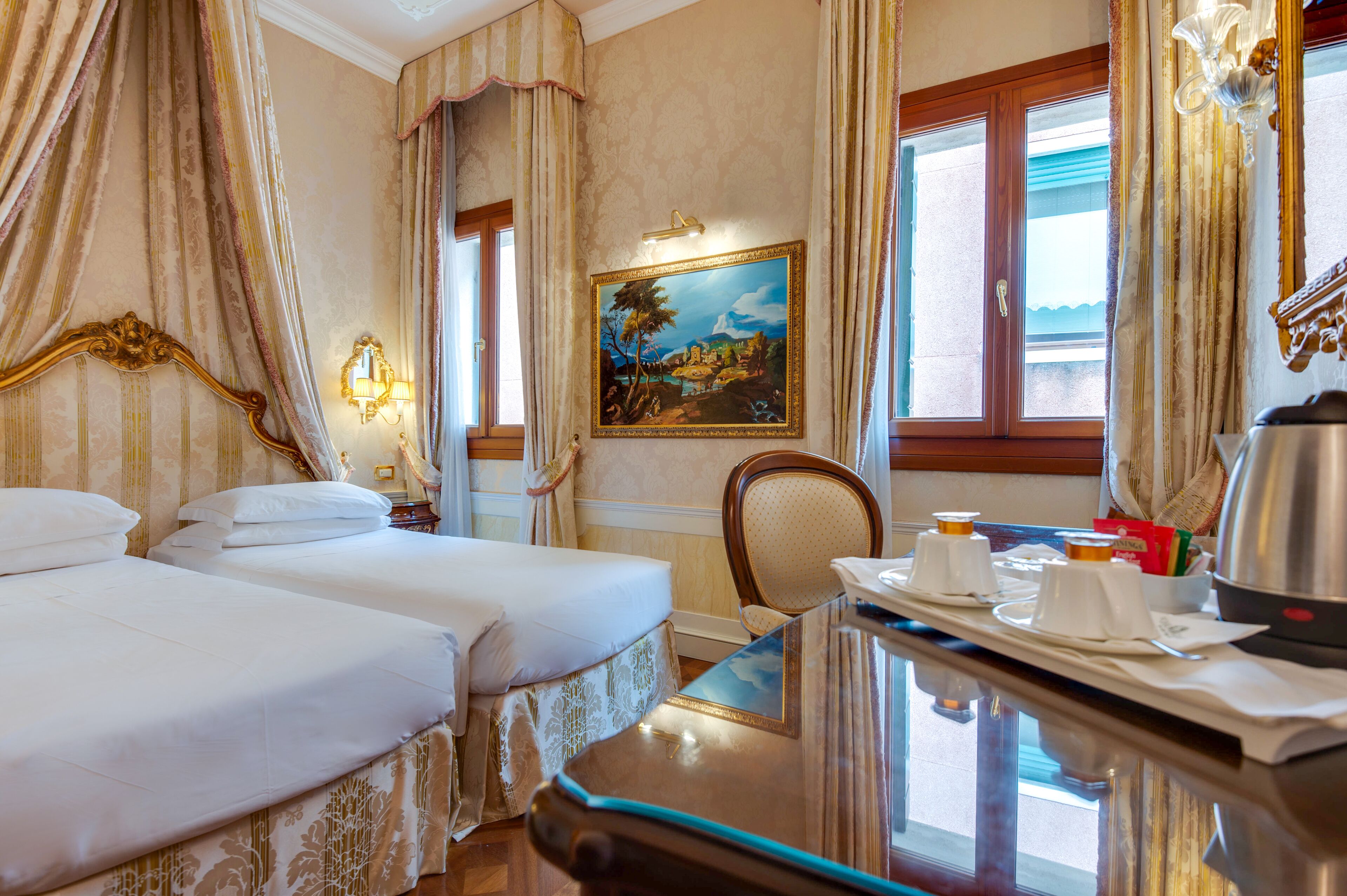 A hotel room with two beds, a painting on the wall, a window with curtains, and a table with a kettle and cups.