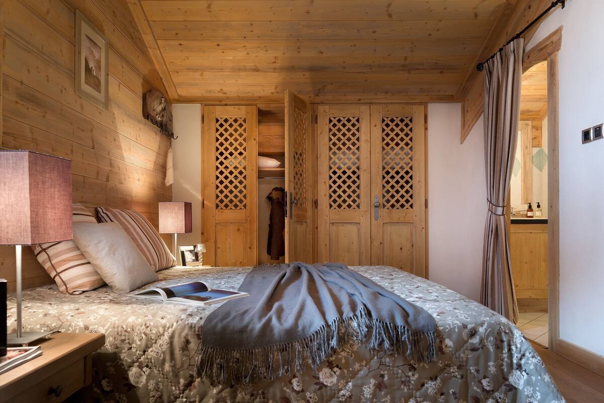 Une pièce chaleureuse aux murs en bois, comprenant un lit, une table de chevet, une lampe et une armoire ornée d’un motif en treillis.