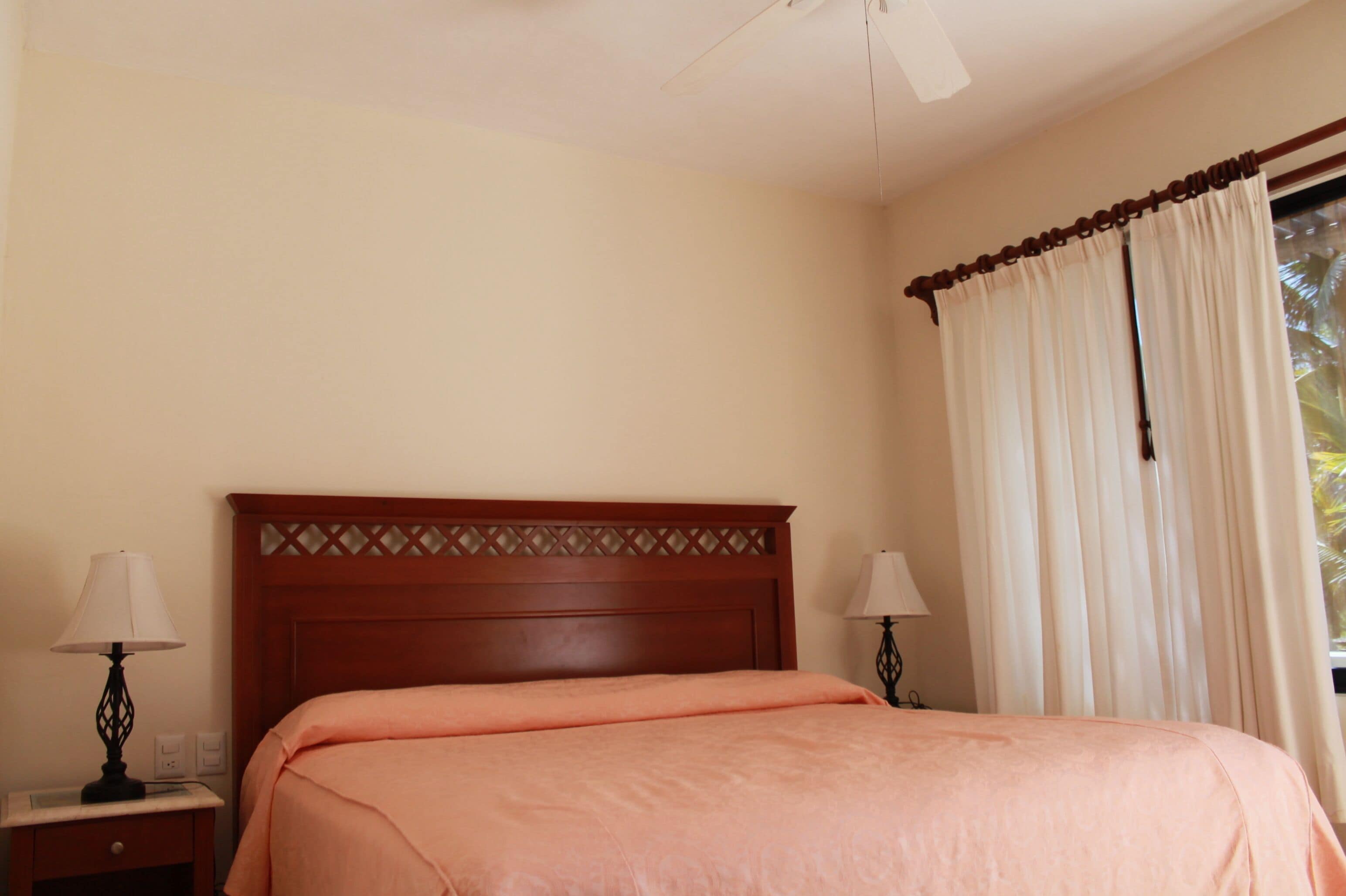 Un dormitorio con cabecera de madera, cama con colcha color melocotón, dos mesitas de noche con lámparas y una ventana con cortinas.