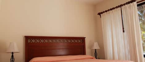 Une chambre à coucher avec une tête de lit en bois, un lit recouvert d’une couvre-lit de couleur pêche, deux tables de chevet avec des lampes et une fenêtre avec des rideaux.