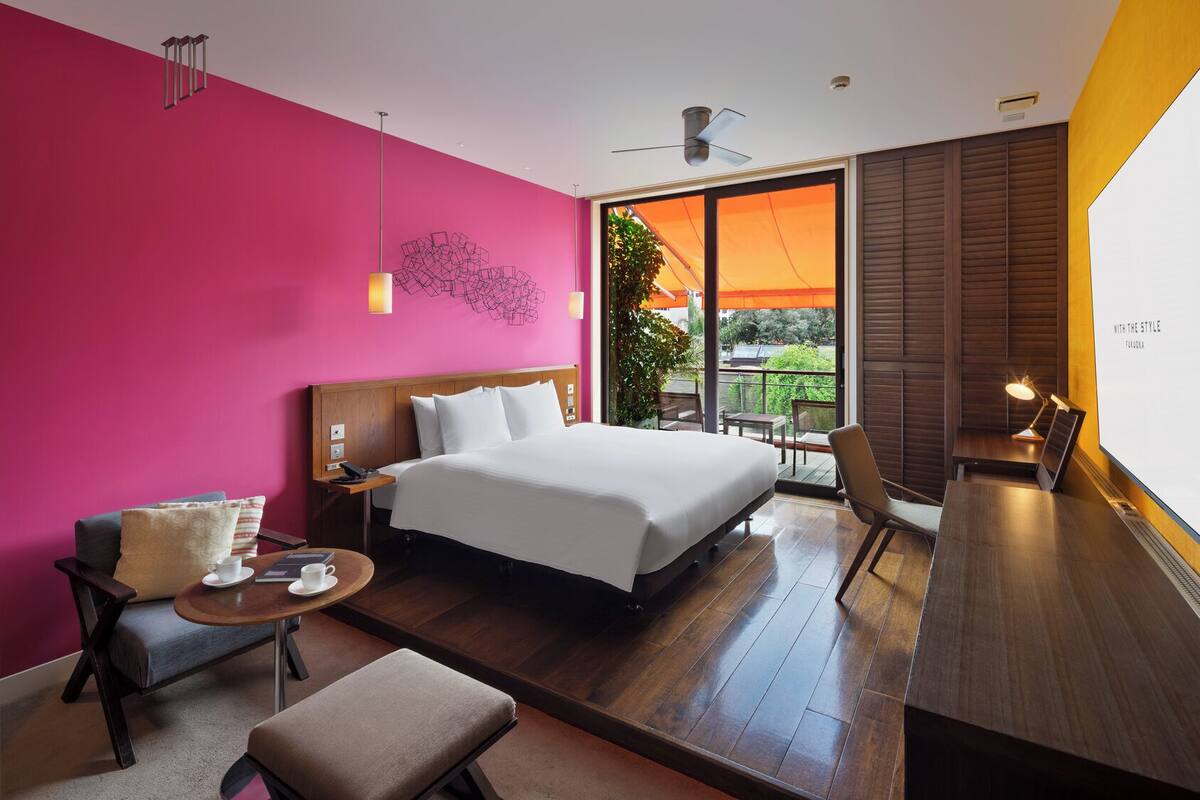 A modern hotel room with a large bed, a desk, a chair, a small table with a cup, and a view of the outdoors.