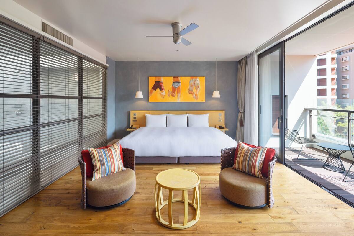 A modern hotel room with a large bed, two armchairs, a small round table, and a balcony with a view of the city.