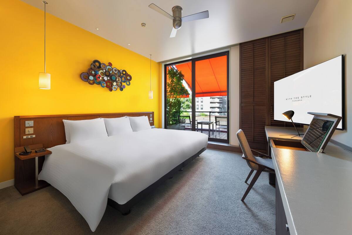 A hotel room with a large bed, a desk with a chair, a ceiling fan, and a view of the outdoors.