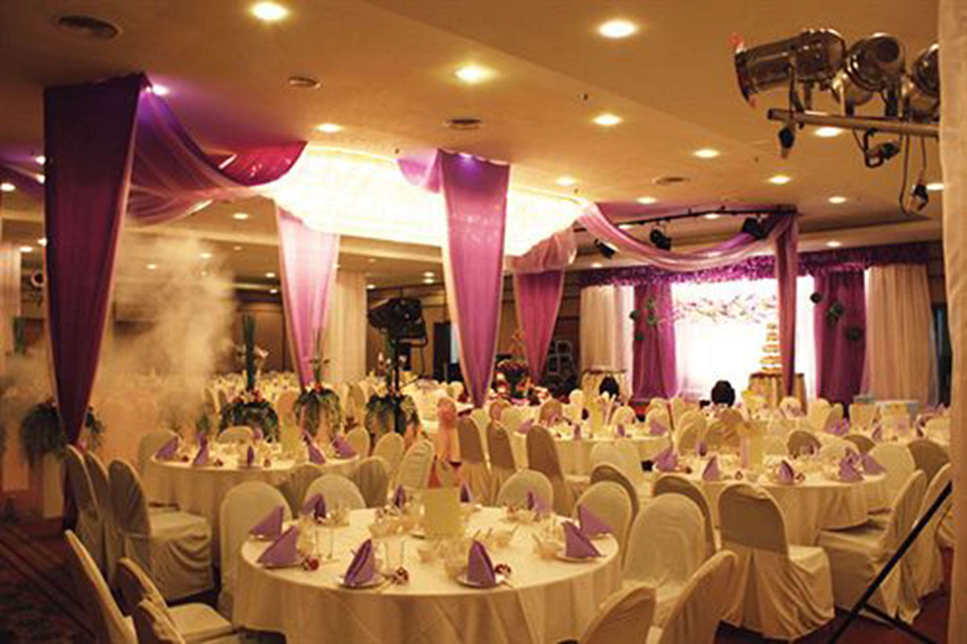 A banquet hall with round tables covered in white tablecloths and purple drapes, equipped with chairs and centerpieces.