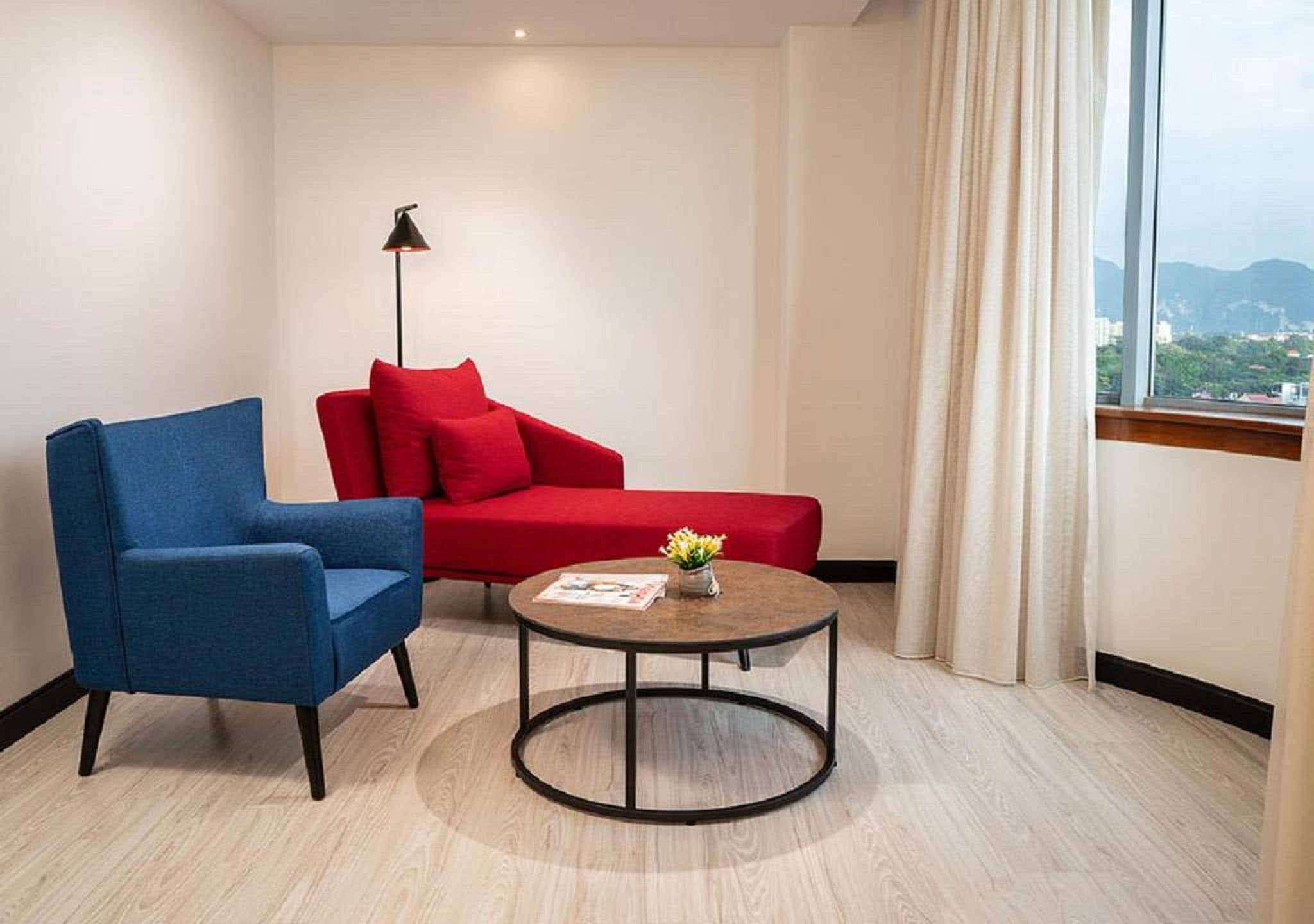 Una habitación con un sillón azul, un sofá rojo, una mesita redonda con una revista y una lámpara de pie.