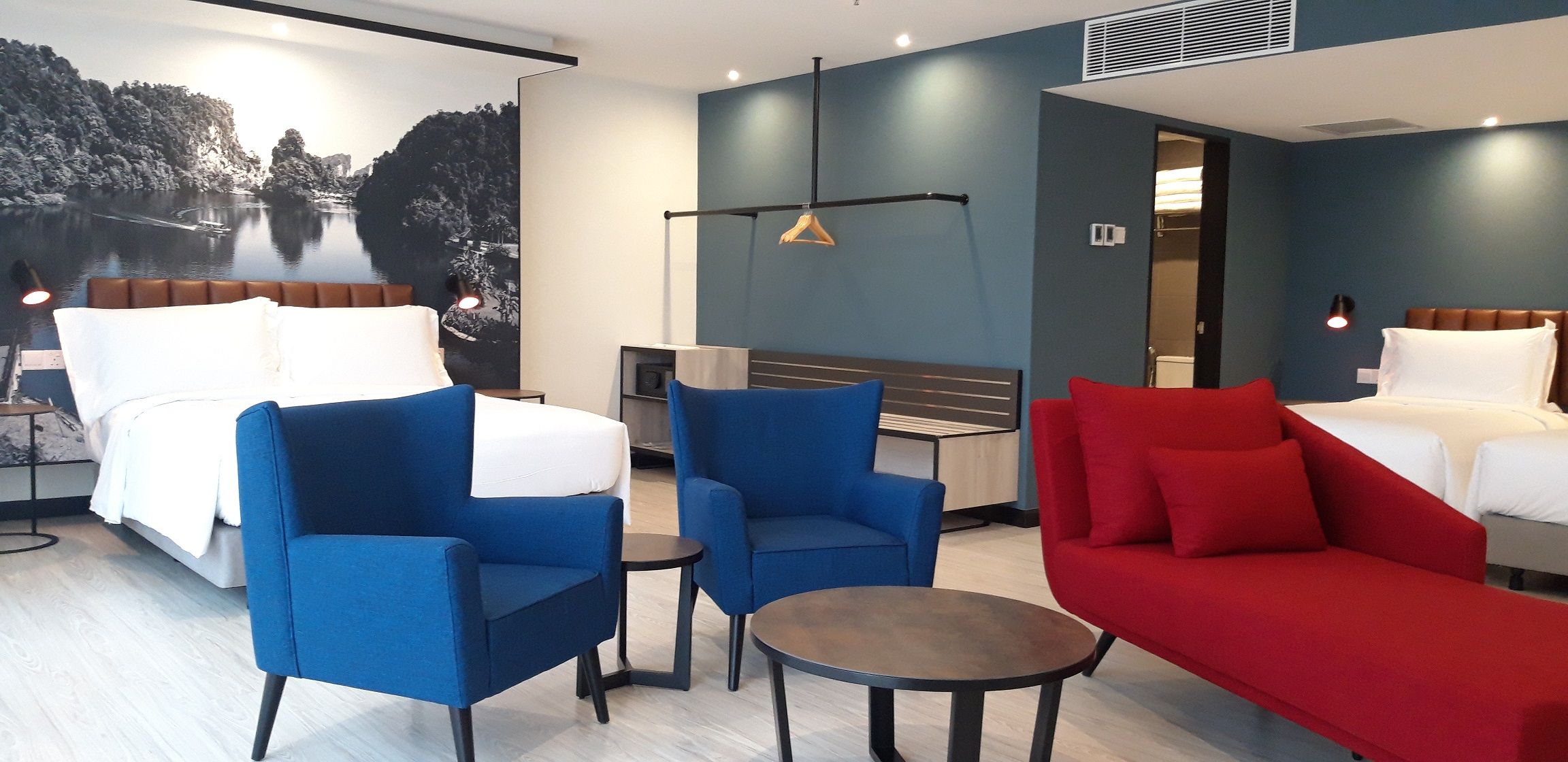 A hotel room with a bed, two blue armchairs, a red sofa, a small round table, and a wall-mounted light fixture.