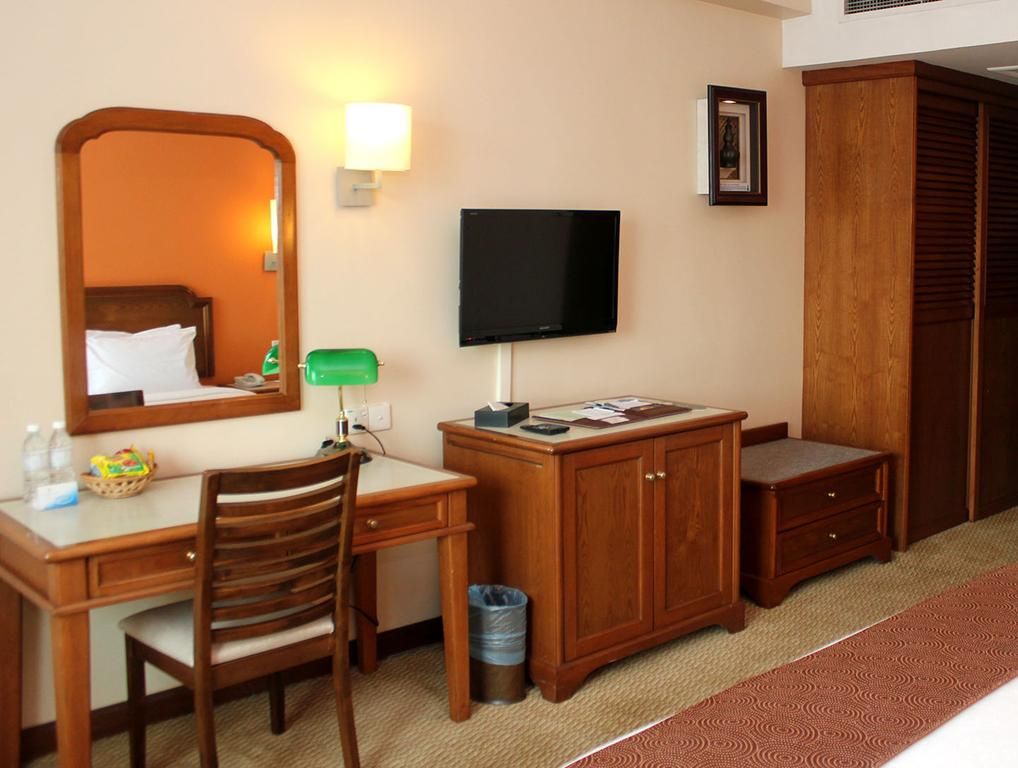 A hotel room with a wooden desk, a chair, a television, a cabinet, a trash bin, and a wardrobe.