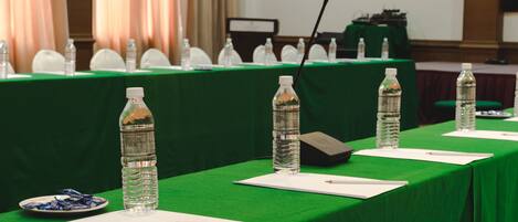 Un salón de conferencias con una larga mesa cubierta con un mantel verde, equipado con botellas de agua, micrófonos y tarjetas de identificación.
