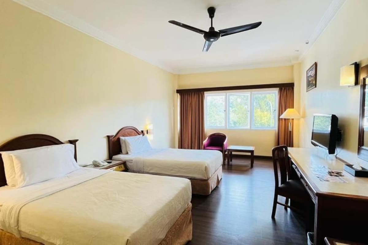 A hotel room with two beds, a desk, a chair, a television, and a ceiling fan.