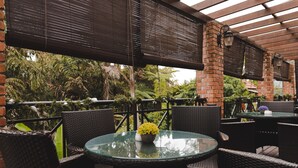 An outdoor patio area with round glass tables and wicker chairs, surrounded by brick walls and covered by a pergola with dark blinds. - Heritage Hotel Cameron Highlands (Tanah Rata)