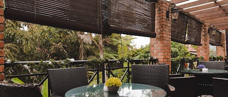 An outdoor patio area with round glass tables and wicker chairs, surrounded by brick walls and covered by a pergola with dark blinds.