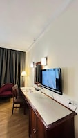 Habitación de hotel con televisión de pantalla plana montada en la pared, un escritorio con silla, lámpara y ventana con cortinas.