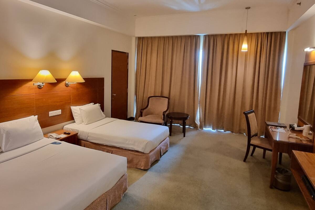 Habitación de hotel con dos camas, un escritorio, una silla, una lámpara y una cortina.