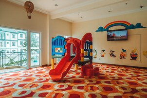 Zona de juegos infantiles con tobogán y estructuras de recreación, alfombra estampada y un amplio ventanal con vistas a edificios.