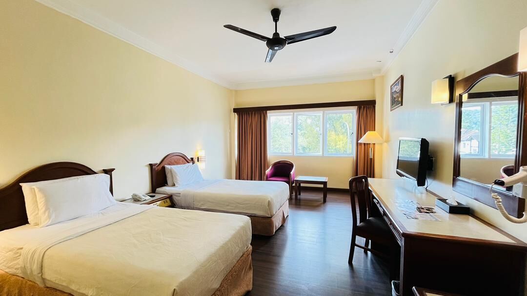 A hotel room with two beds, a desk, a chair, a television, and a ceiling fan.