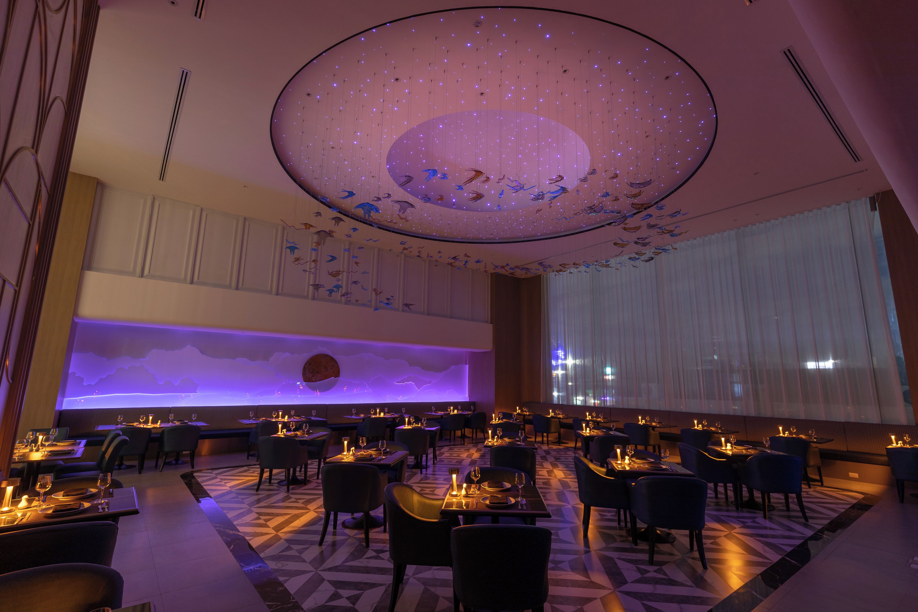 Un restaurant spacieux doté d’un plafond haut, éclairé par un luminaire circulaire original. Les tables sont disposées avec soin et agrémentées de bougies, et une grande fenêtre offre une vue sur le paysage urbain.