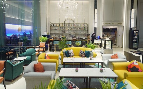 A modern hotel lobby with a central seating area, a chandelier, and a visible restaurant named "Crystal Restaurant" in the background.